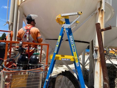 Worker using an access platform and ladder beside a large industrial vessel while wearing fall protection.